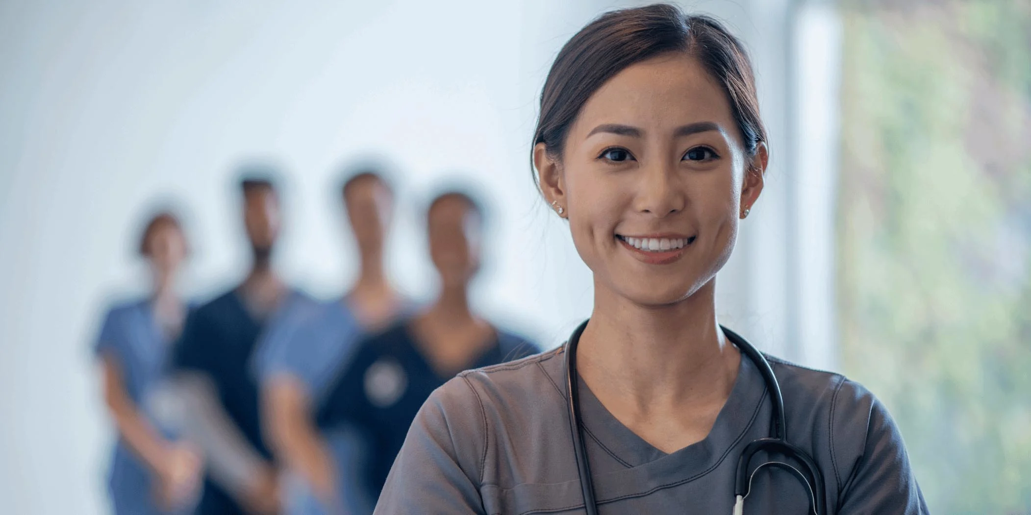 An Asian woman nurse smiles as she stands in front of her team in the hospital hallway.